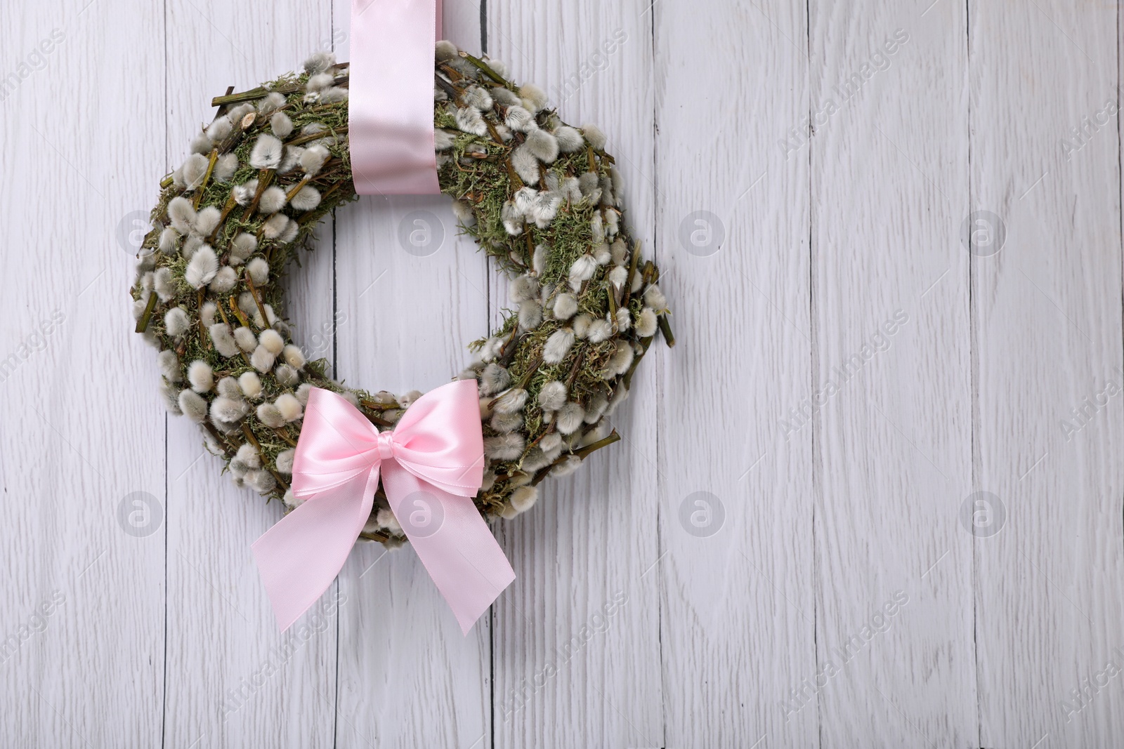 Photo of Wreath made of beautiful willow branches and pink bow hanging on white wooden background. Space for text