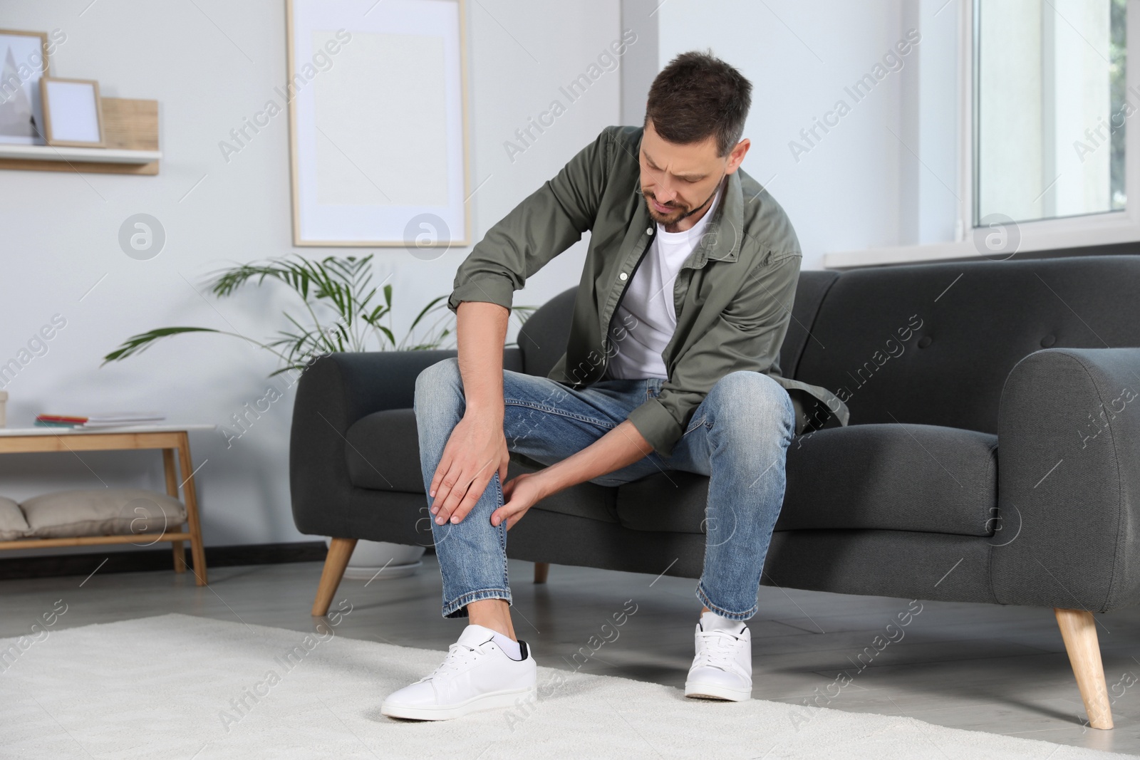 Photo of Man suffering from pain in leg on sofa at home