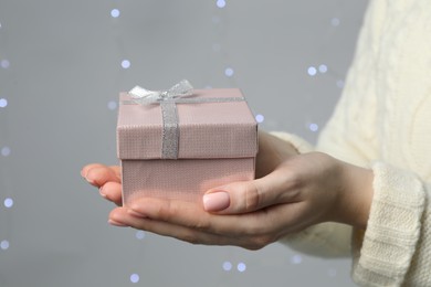 Photo of Christmas present. Woman holding gift box against grey background with blurred lights, closeup