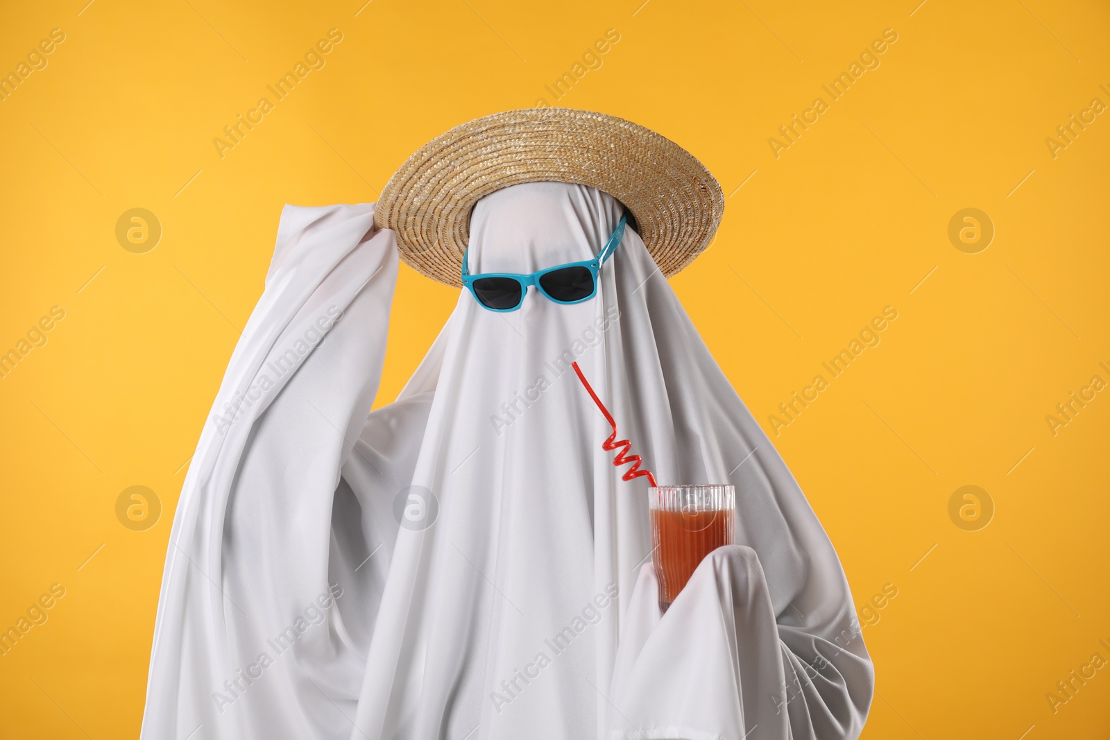 Photo of Person in ghost costume, sunglasses and straw hat holding glass of drink on yellow background