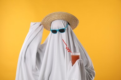 Photo of Person in ghost costume, sunglasses and straw hat holding glass of drink on yellow background