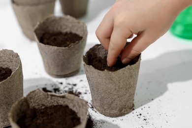 Little girl planting vegetable seeds into peat pots with soil at white table, closeup