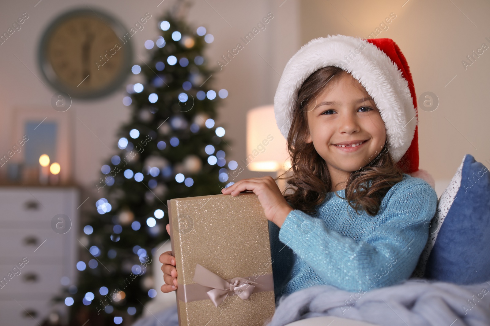 Photo of Cute little child in Santa hat with Christmas gift box sitting on sofa at home