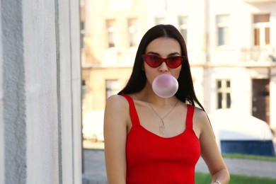 Photo of Beautiful woman in stylish sunglasses blowing gum near building outdoors, space for text