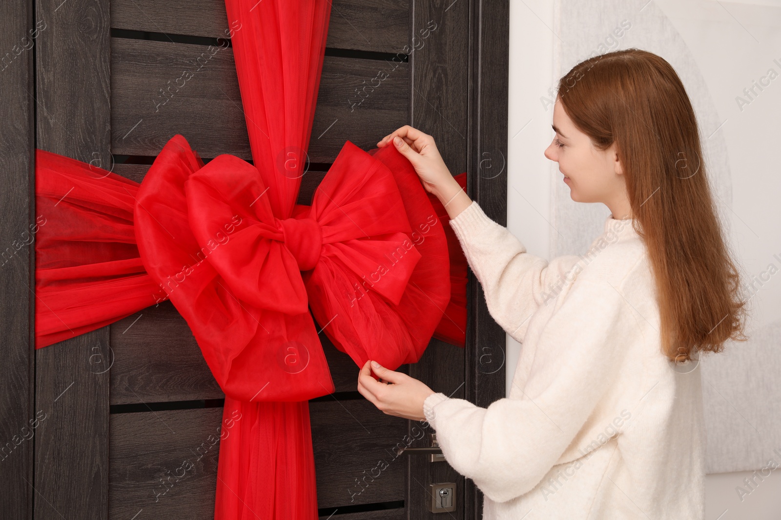 Photo of Woman decorating wooden door with red bow indoors. Christmas mood