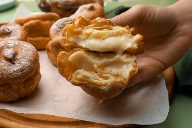 Photo of Woman holding delicious profiterole filled with cream above green table, closeup