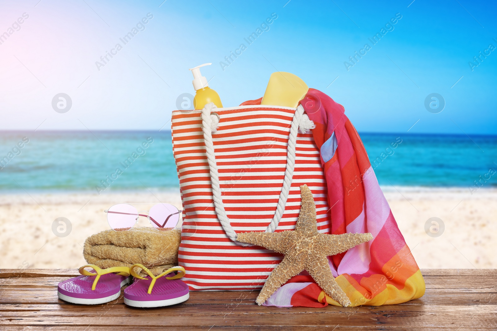 Image of Stylish striped bag and other beach accessories on wooden table near sea