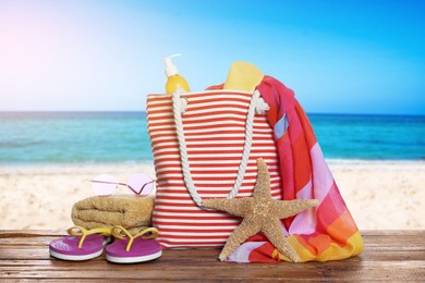 Image of Stylish striped bag and other beach accessories on wooden table near sea