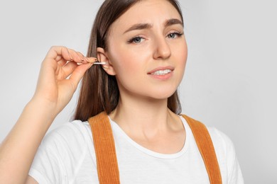 Young woman cleaning ear with cotton swab on light grey background, closeup