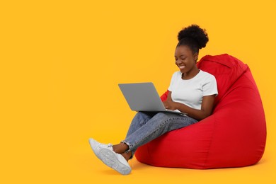 Beautiful young woman with laptop sitting on beanbag chair against orange background. Space for text