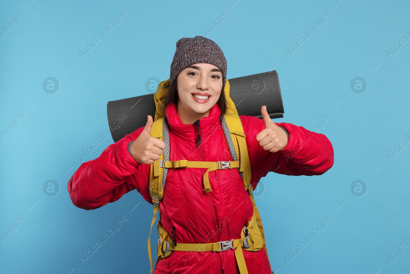 Photo of Smiling young woman with backpack showing thumbs up on light blue background. Active tourism