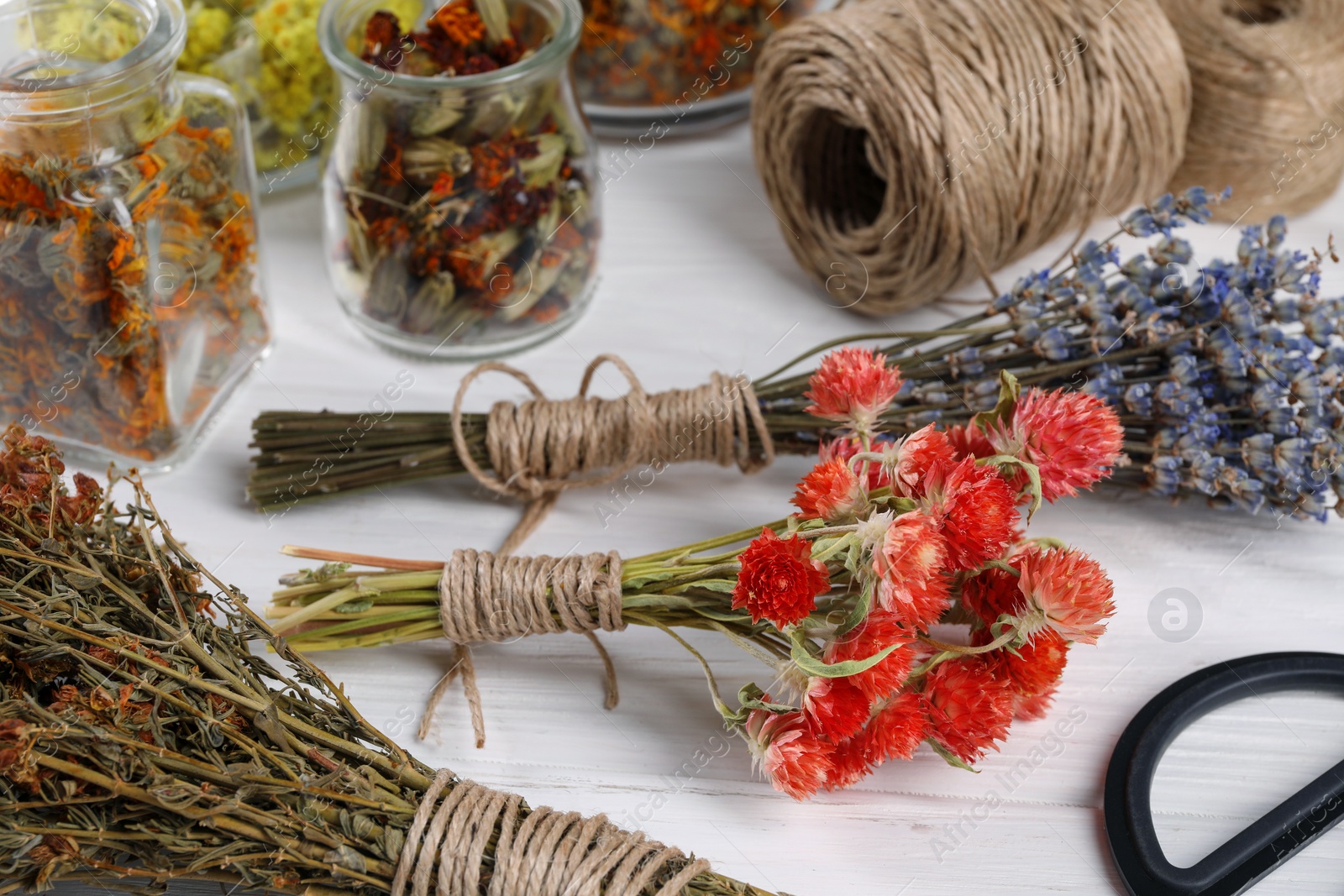 Photo of Bunches of dry flowers, different medicinal herbs and spools on white wooden table