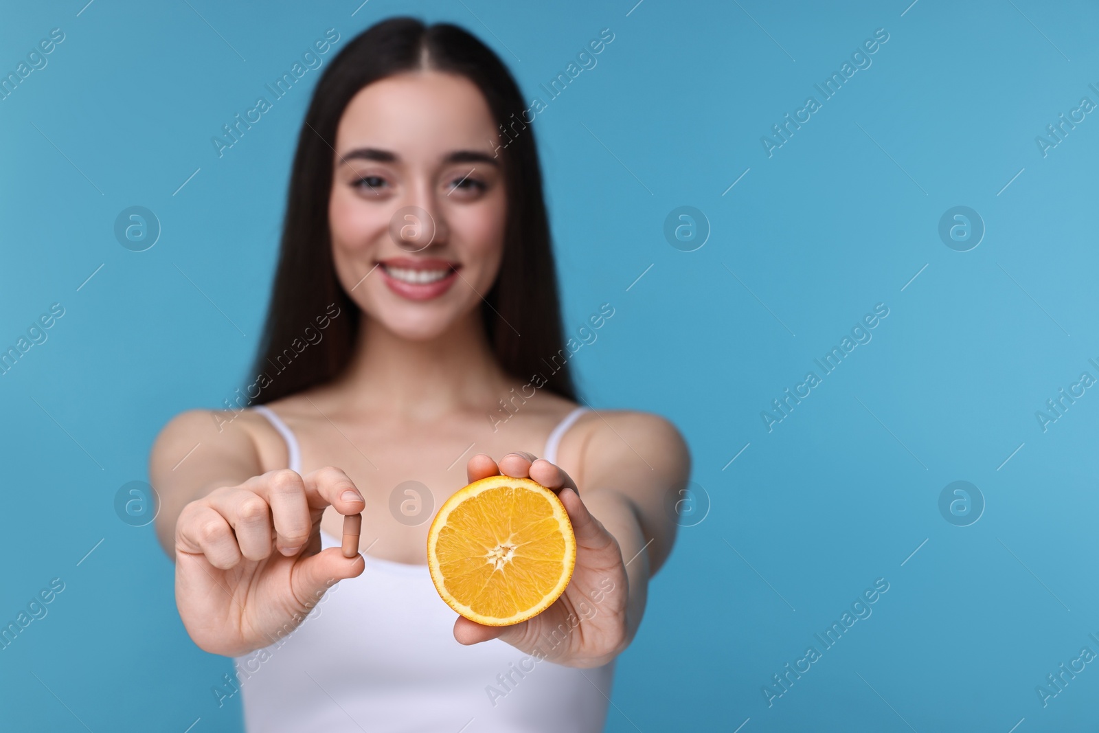 Photo of Beautiful young woman with vitamin pill and piece of orange on light blue background, selective focus. Space for text