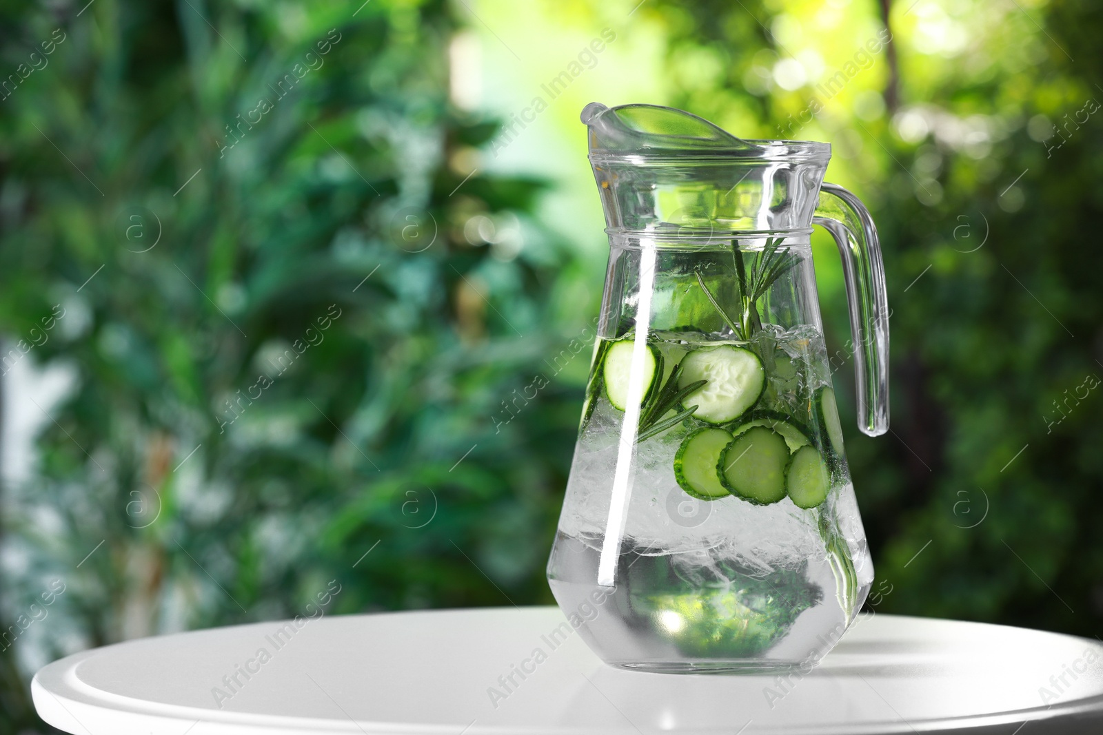 Photo of Refreshing cucumber water with rosemary in jug on white table against blurred green background. Space for text