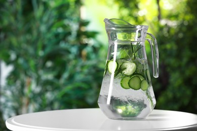 Refreshing cucumber water with rosemary in jug on white table against blurred green background. Space for text