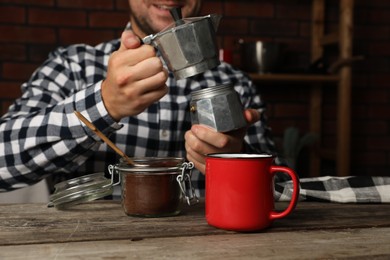 Photo of Man with moka pot in kitchen, focus on jar of ground coffee and mug on wooden table