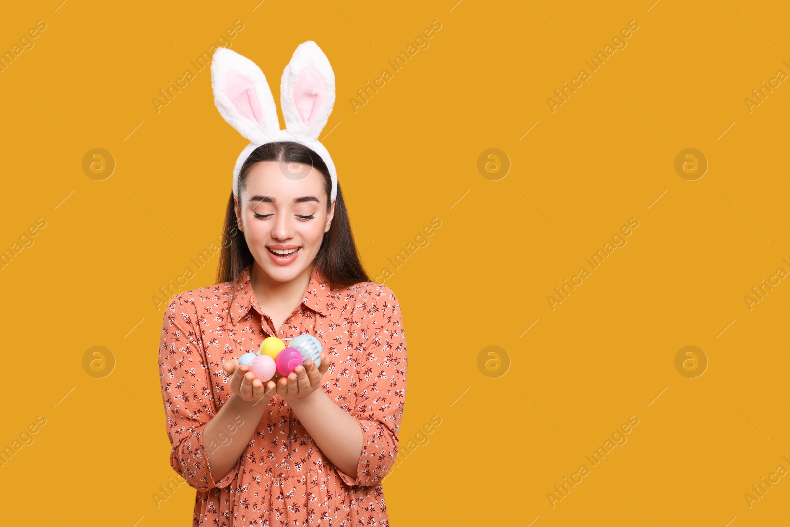 Photo of Happy woman in bunny ears headband holding painted Easter eggs on orange background. Space for text.