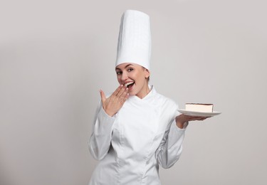 Happy professional confectioner in uniform holding delicious cheesecake on light grey background