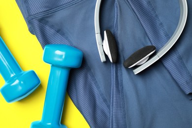 Photo of Stylish sports leggings, headphones and dumbbells on yellow background, flat lay