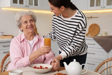 Young caregiver serving breakfast for senior woman at table in kitchen. Home care service