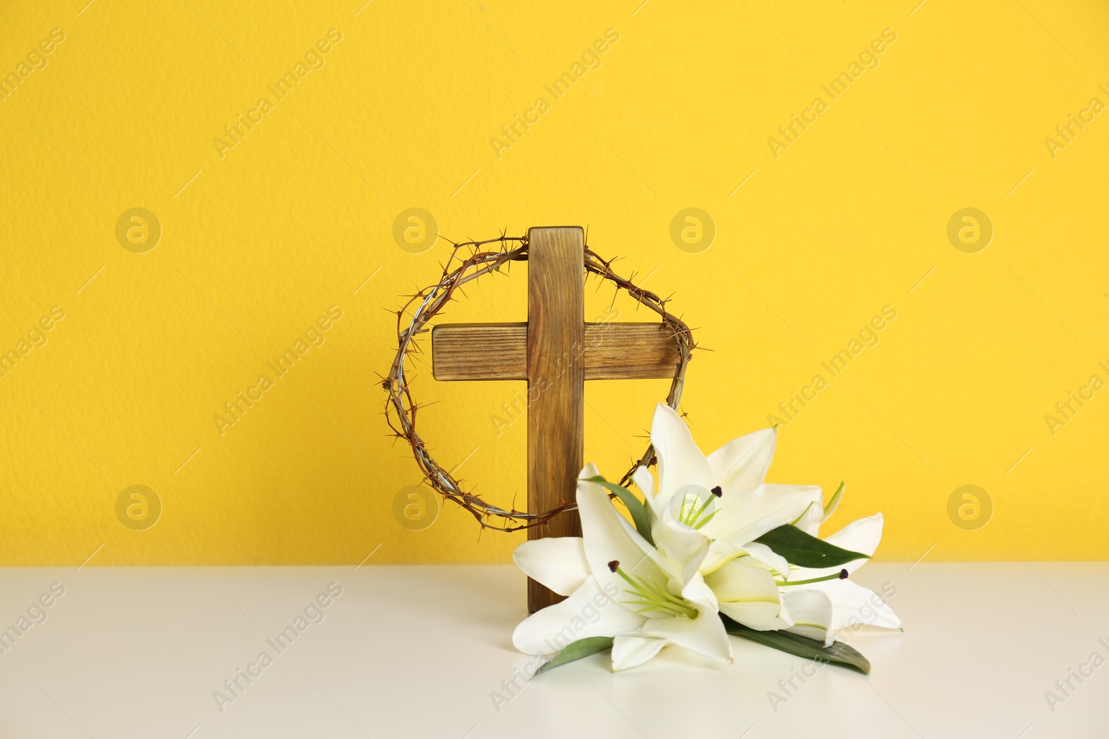 Photo of Wooden cross, crown of thorns and blossom lilies on table against color background