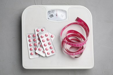 Photo of Scales with weight loss pills and measuring tape on grey table, top view