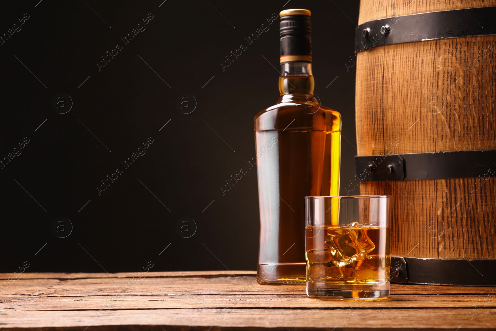 Photo of Whiskey with ice cubes in glass, bottle and barrel on wooden table against black background, space for text