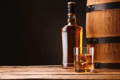 Whiskey with ice cubes in glass, bottle and barrel on wooden table against black background, space for text