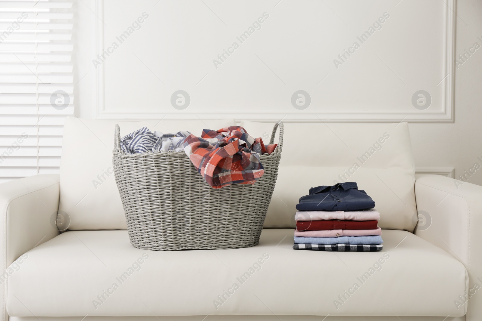 Photo of Wicker basket with dirty laundry and stack of clean clothes on sofa indoors