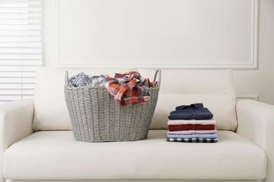 Photo of Wicker basket with dirty laundry and stack of clean clothes on sofa indoors