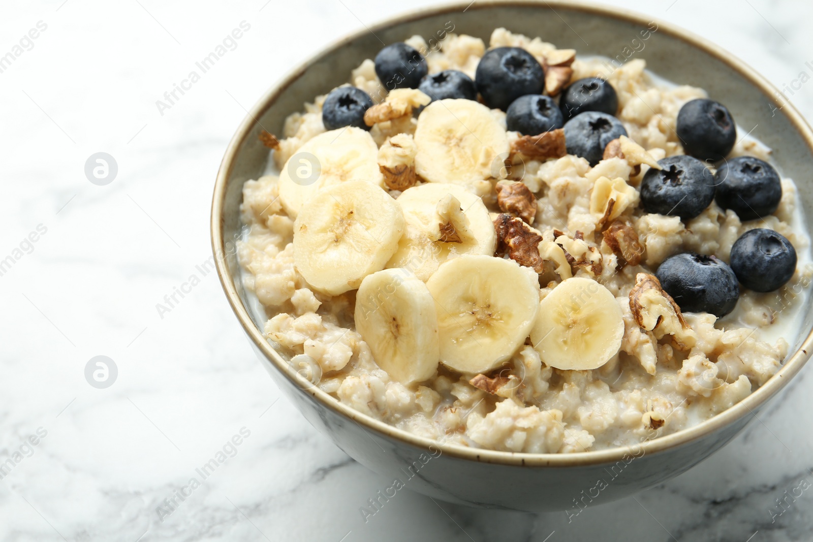 Photo of Tasty oatmeal with banana, blueberries, walnuts and milk served in bowl on white marble table, closeup. Space for text