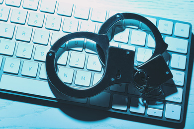 Photo of Handcuffs and computer keyboard on table, top view. Cyber crime