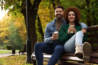 Happy young couple with cups of coffee spending time together on wooden bench in autumn park, space for text