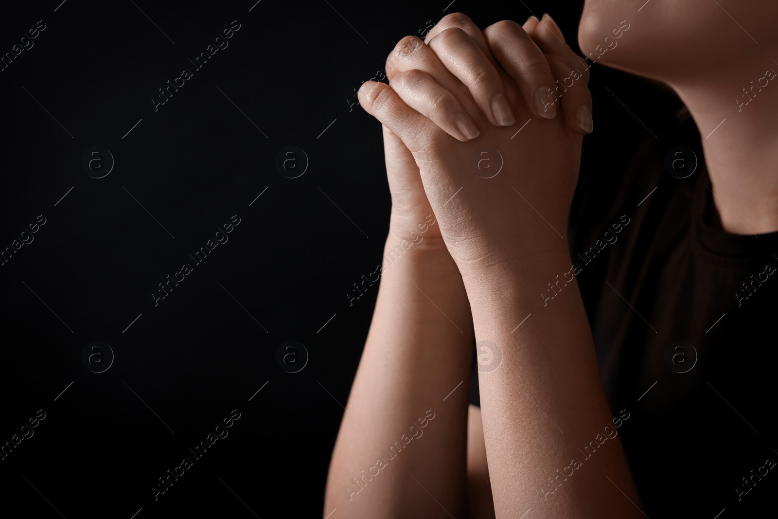 Photo of Woman holding hands clasped while praying against black background, closeup. Space for text