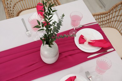 Photo of Table setting. Glasses of tasty beverage, plates, pink napkins and vase with green branches in dining room, above view