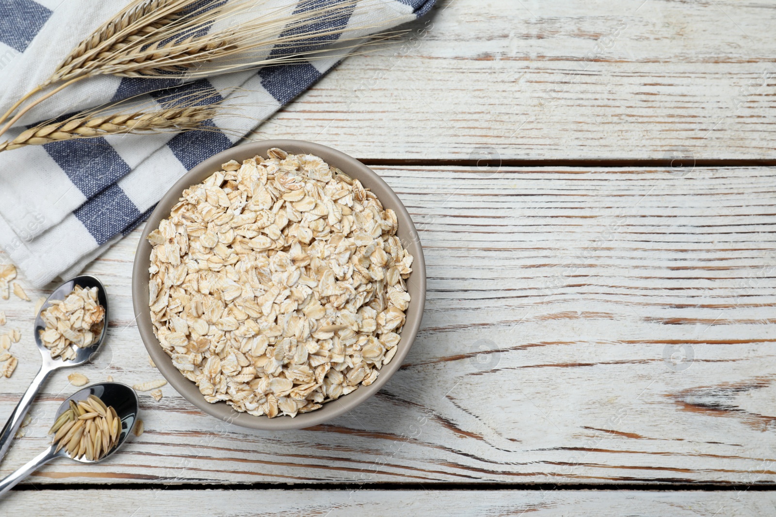 Photo of Bowl of oatmeal, spoons, napkin and spikelets on white wooden table, flat lay. Space for text