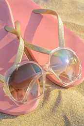 Stylish sunglasses and pink flip flops on sandy beach, closeup