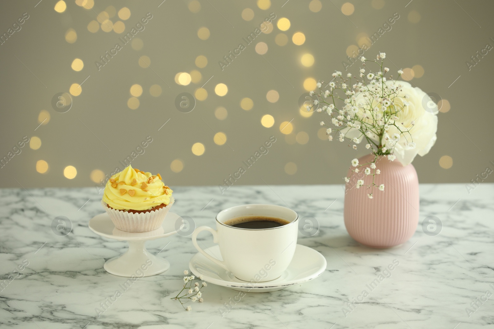 Photo of Delicious cupcake with yellow cream, coffee and flowers on white marble table against blurred lights