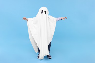 Child in white ghost costume on light blue background. Halloween celebration