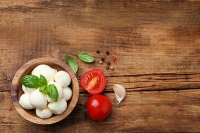 Photo of Delicious mozzarella balls in bowl, tomatoes and basil leaves on wooden table, flat lay. Space for text