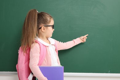 Photo of Happy little school child with notebooks pointing at chalkboard. Space for text