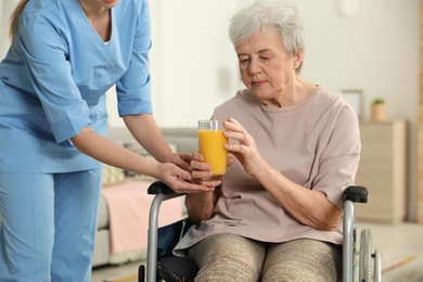 Photo of Nurse giving glass of juice to elderly woman in wheelchair indoors. Assisting senior people
