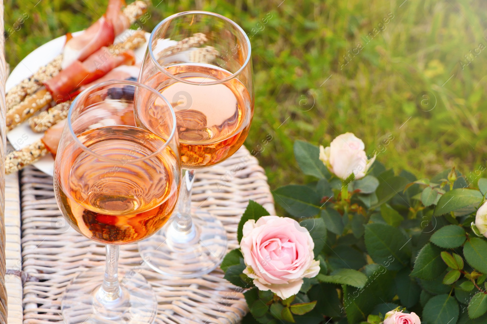 Photo of Flowers near glasses of delicious rose wine and food on picnic basket outdoors, closeup