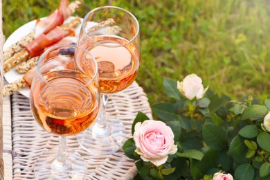 Photo of Flowers near glasses of delicious rose wine and food on picnic basket outdoors, closeup