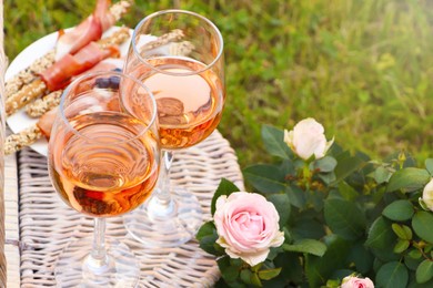 Flowers near glasses of delicious rose wine and food on picnic basket outdoors, closeup