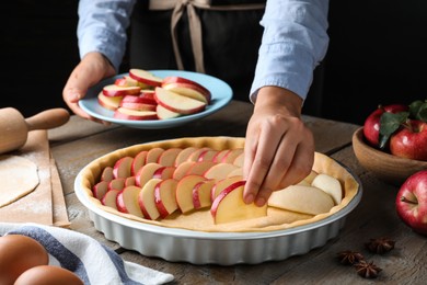 Photo of Woman putting apple slices into dish with raw dough at wooden table, closeup. Baking pie