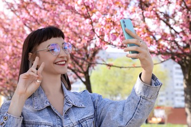 Beautiful young woman taking selfie in park with blossoming sakura trees