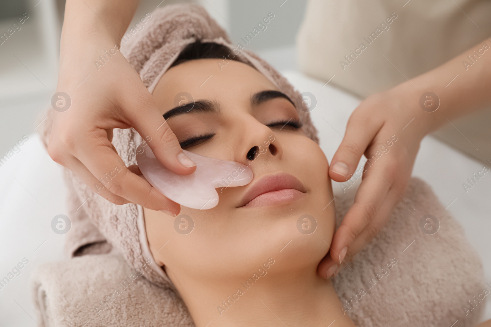 Photo of Young woman receiving facial massage with gua sha tool in beauty salon