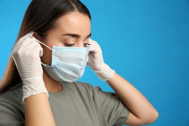 Woman in medical gloves putting on protective face mask against blue background