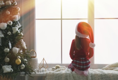 Photo of Cute little girl in Santa hat on window sill near Christmas tree at home, back view